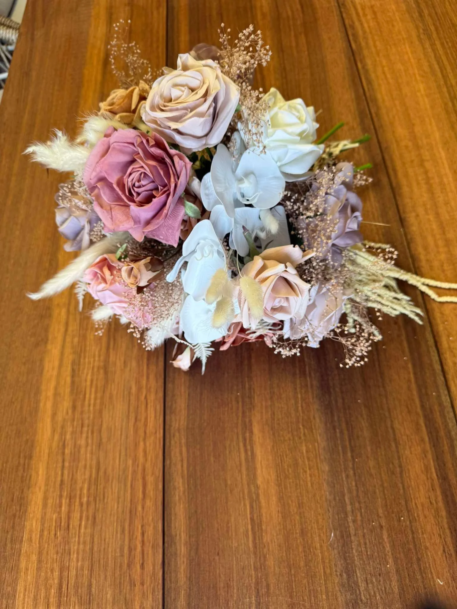 A floral arrangement featuring light pink, white, and cream roses, complemented by dried plants and greenery, placed on a wooden surface.