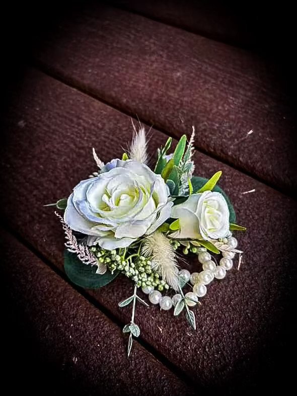 A white floral boutonniere with white roses, green leaves, and decorative pearls on a dark brown textured wooden surface.