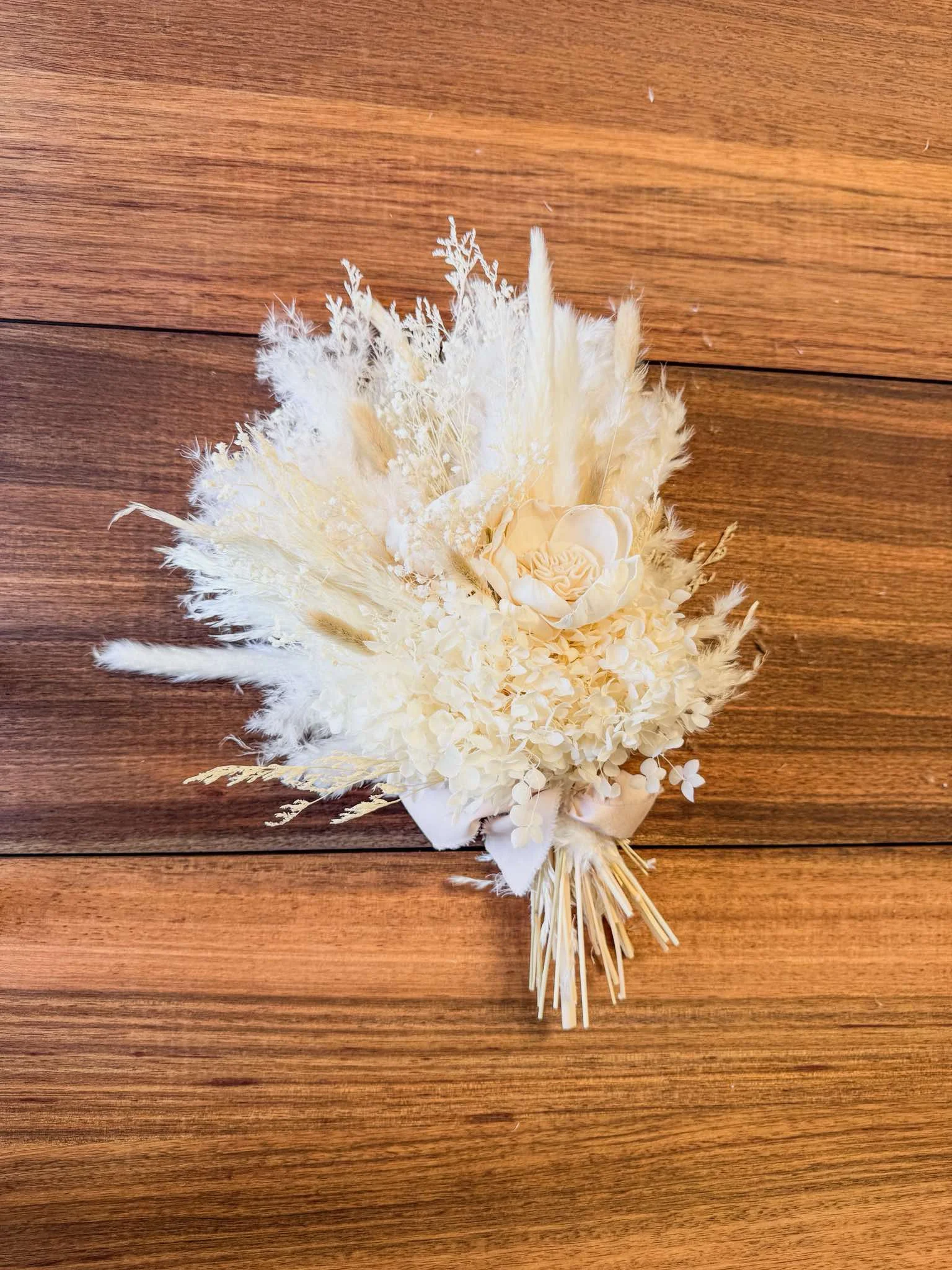 Cream-colored flower bouquet with pampas grass and hydrangeas, tied with a cream ribbon, placed on a wooden surface.