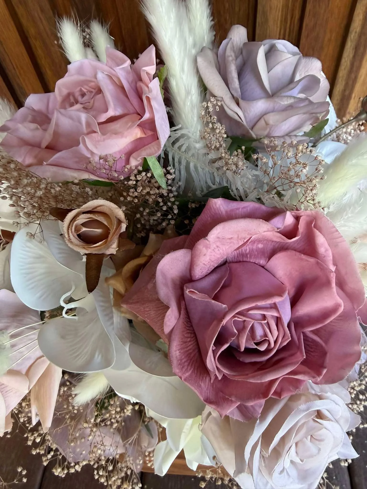 A floral arrangement featuring pink and white roses, white orchids, dried baby's breath, and various filler flowers and greenery, set against a wooden background.
