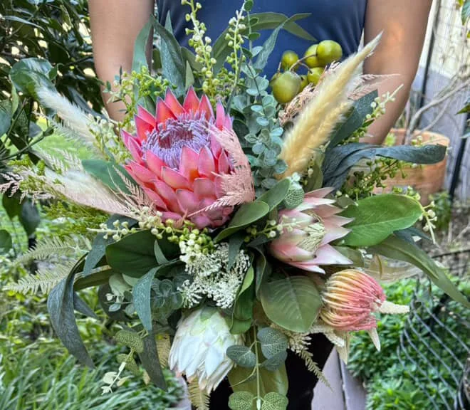 Arranged bouquet of tropical and exotic flowers including protea, with green foliage, in a garden setting.