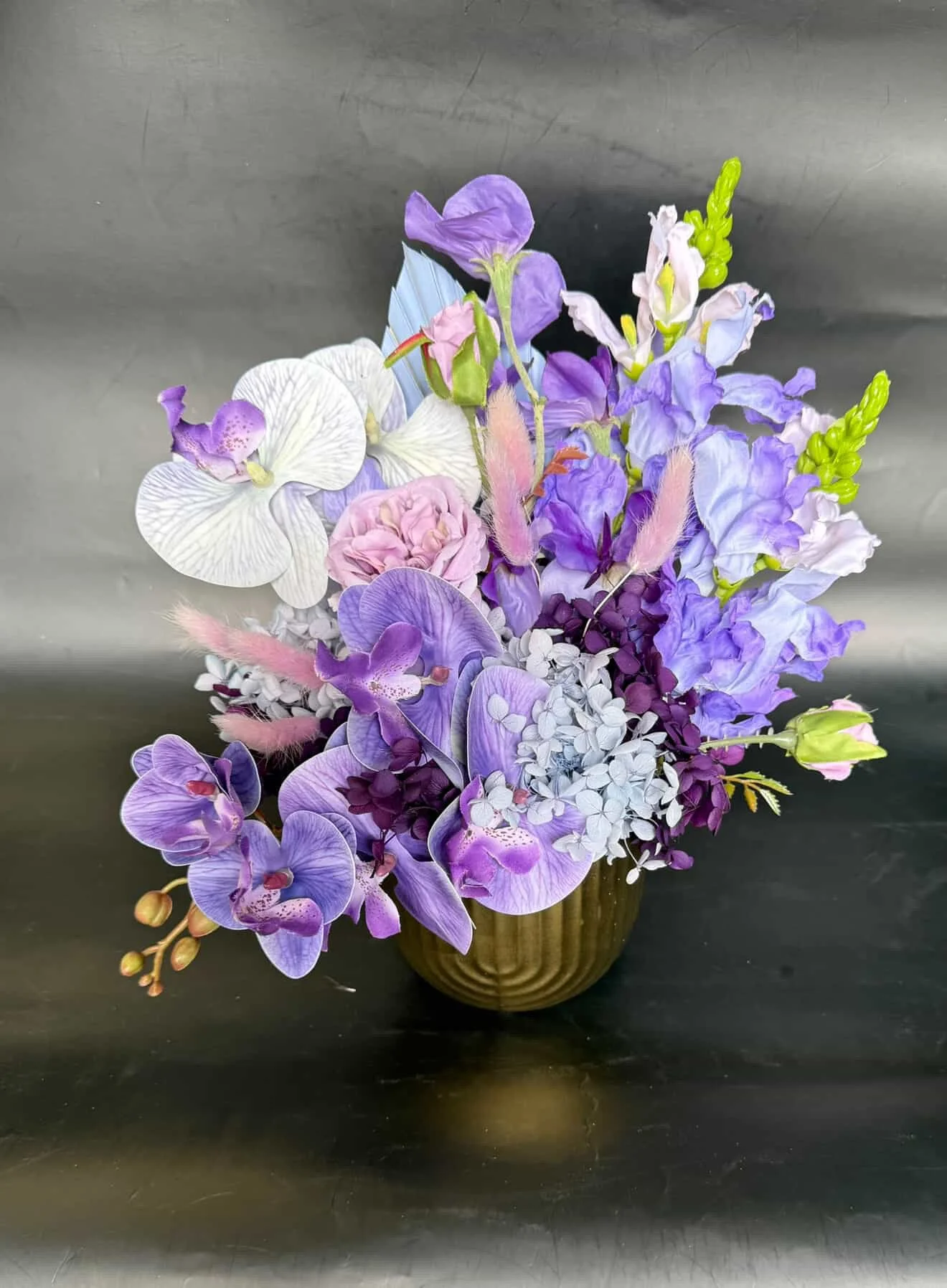 A colorful bouquet of purple, white, and pink flowers in a brown vase, set against a metallic gray background.