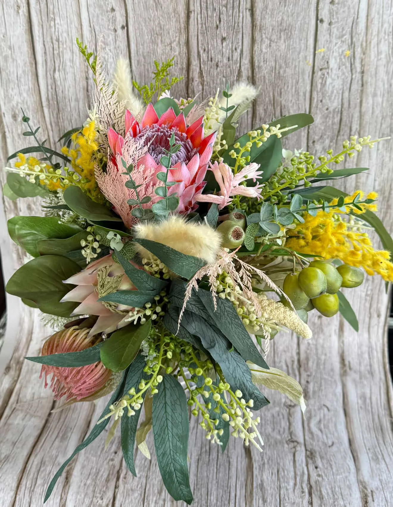 A colorful bouquet of flowers in a basket on a wooden background, featuring pink protea, yellow blossoms, greenery, and berries.