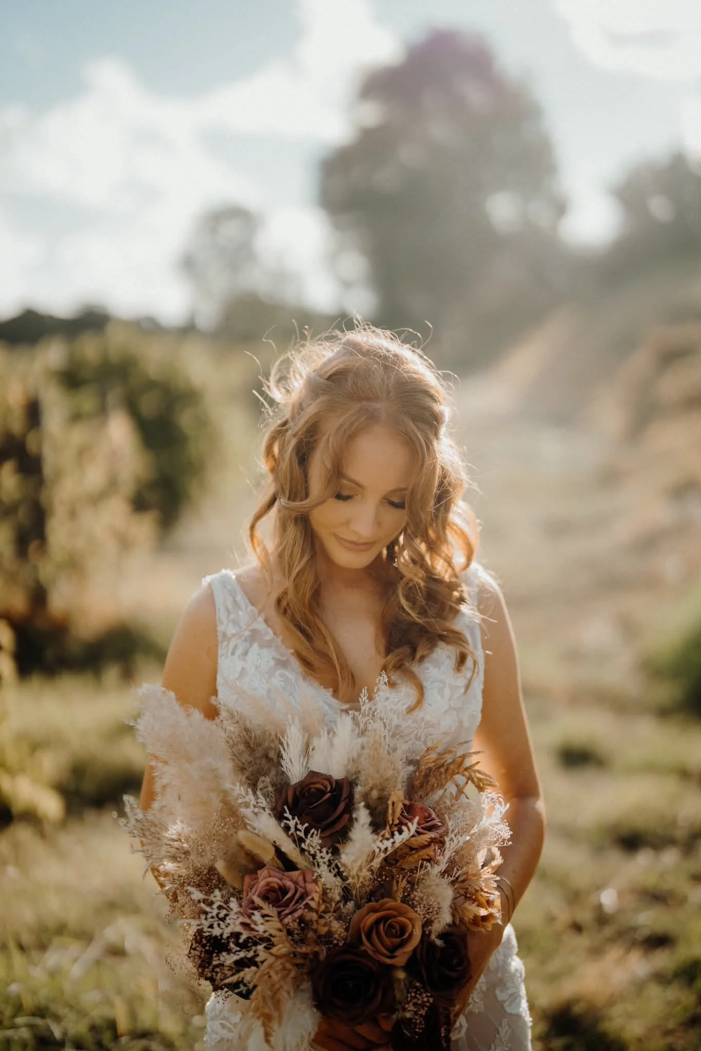 A woman with long, curly, red hair wearing a white lace dress, standing outdoors, holding a bouquet of brown and beige roses and dried flowers, with trees and a sky with clouds in the background.
