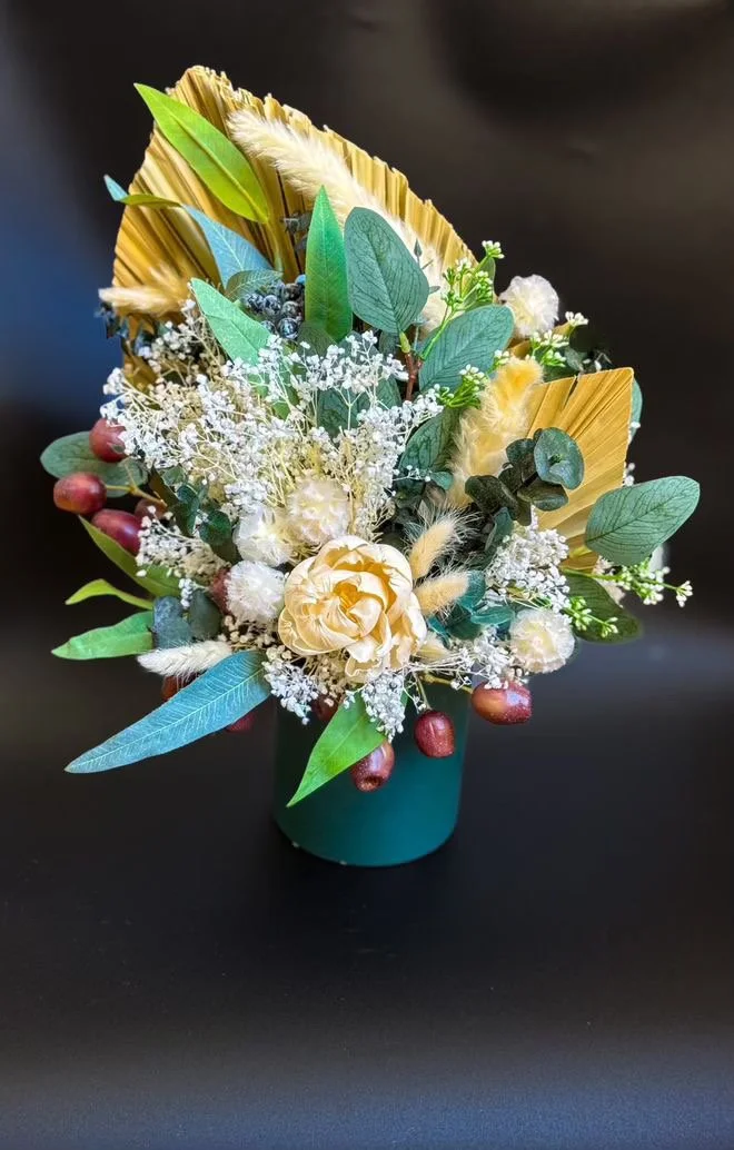 A decorative floral arrangement in a teal container featuring white roses, baby's breath, green leaves, dried palm leaves, and sprigs of red berries, set against a black background.