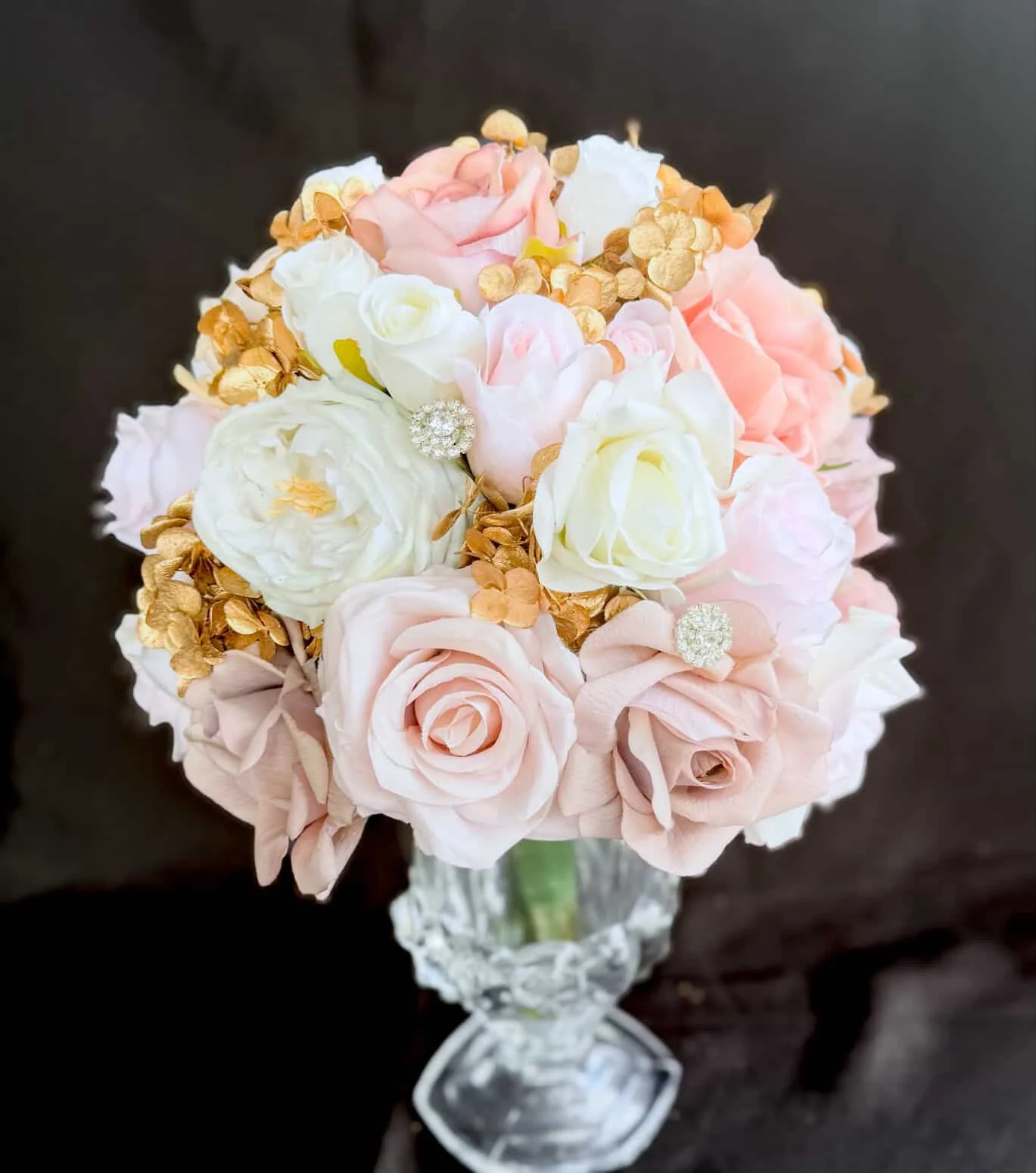 A bouquet of pink, white, and cream roses with gold-dusted dried hydrangea petals and decorative rhinestone accents, arranged in a glass vase with a faceted design.