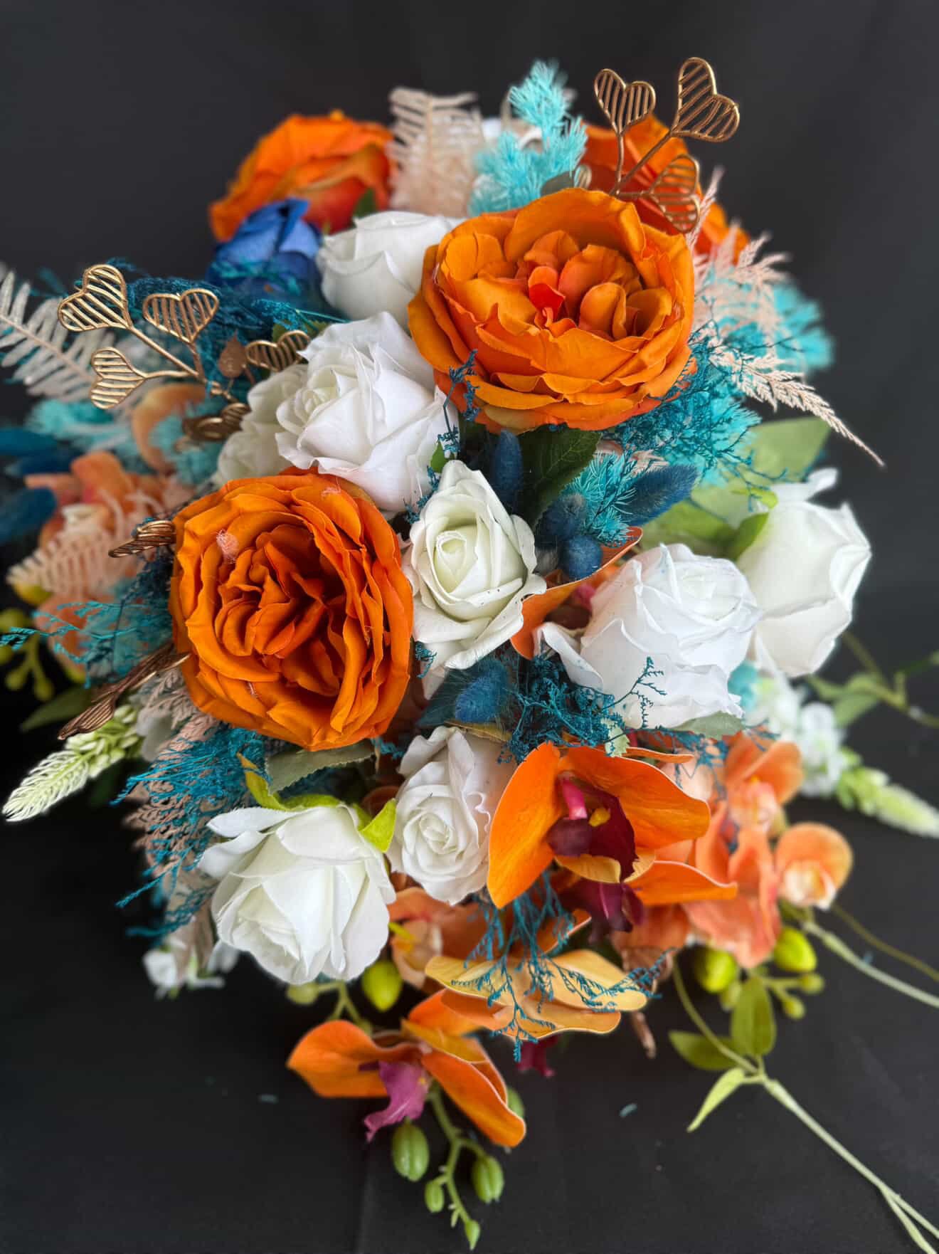 Colorful artificial flower bouquet featuring orange and white roses, orange orchids, blue feathers, gold heart-shaped decorative picks, and other greenery against a black background.