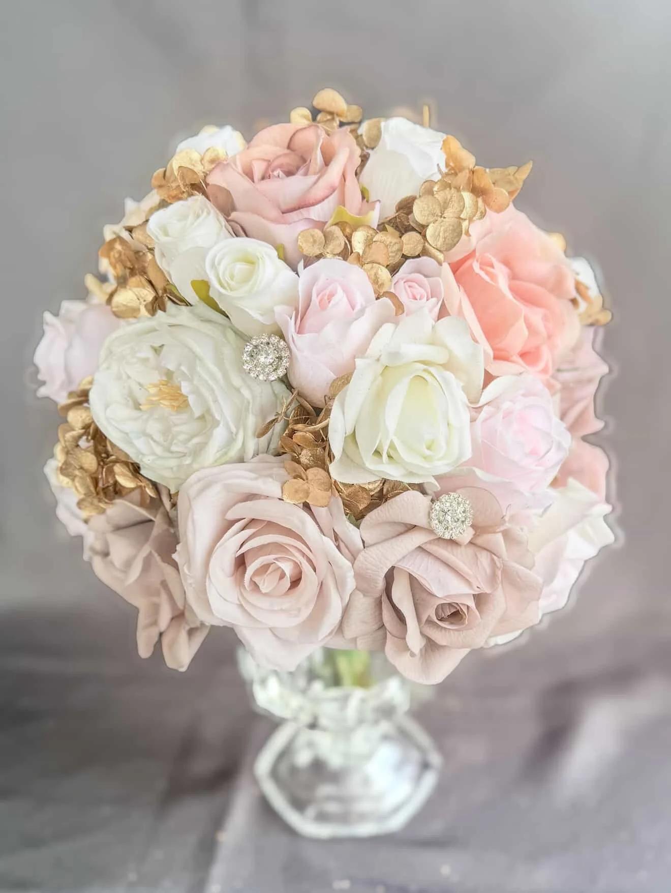 A floral bouquet featuring white and blush roses, with gold and pearl embellishments, displayed in a glass vase against a gray background.