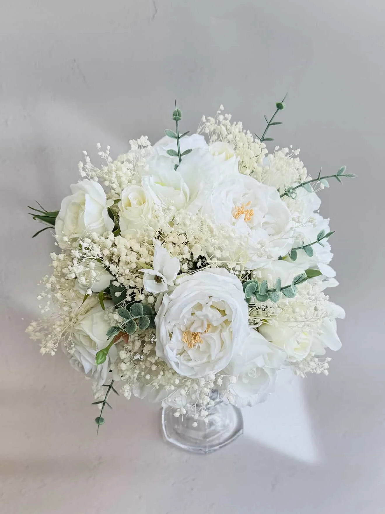 A white floral bouquet in a clear glass vase, featuring white roses, peonies, baby's breath, and eucalyptus leaves on a light background.