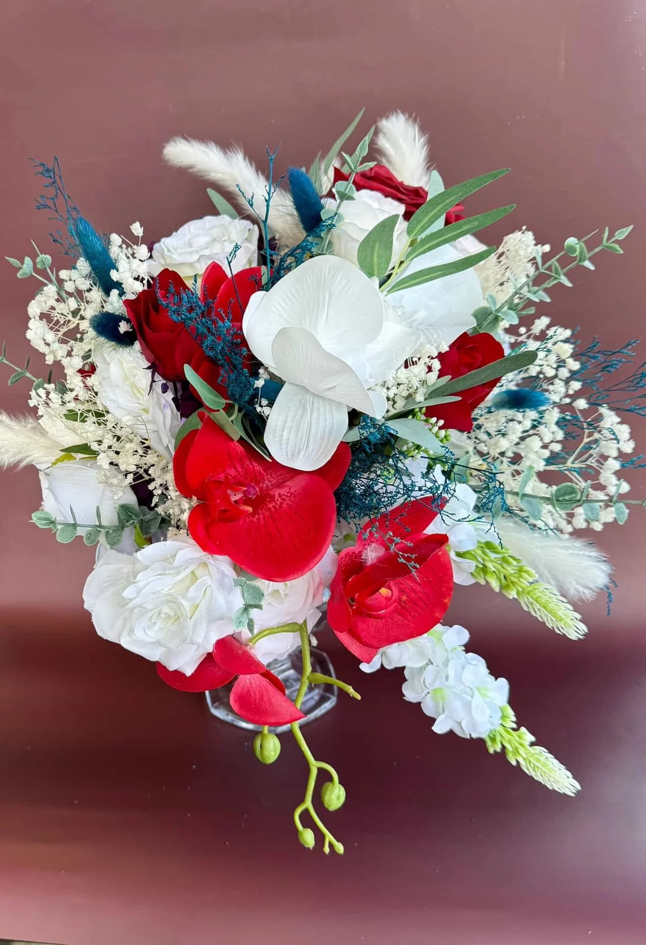 A colorful bouquet of white, red, and blue flowers with green foliage in a glass vase on a brown surface.