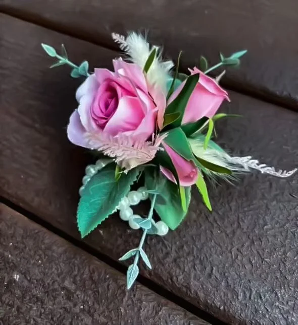 A boutonniere with a pink rose, pink calla lily, green leaves, white feathers, and a strand of white beads on a dark wooden surface.