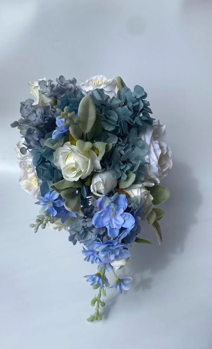 A bouquet of artificial flowers with white, blue, and green blooms and leaves, set against a plain light background.