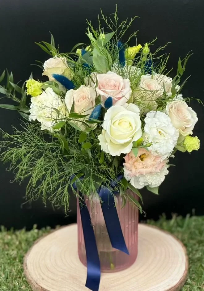 A floral arrangement with white and light pink roses, green foliage, and blue accents in a pink glass vase, placed on a wooden surface against a dark background.