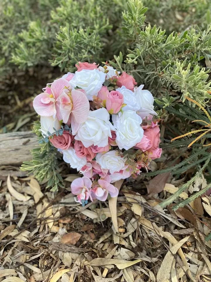 A bouquet of pink and white flowers, including roses and orchids, placed on dry leaves and green plants outdoors.