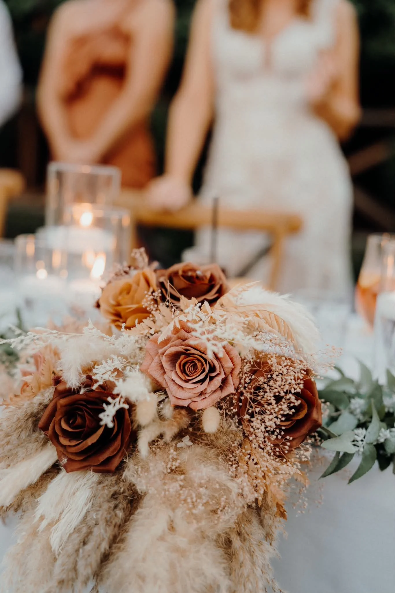 Close-up of a floral arrangement with brown and peach roses, white feathers, and filler flowers on a furry beige surface.