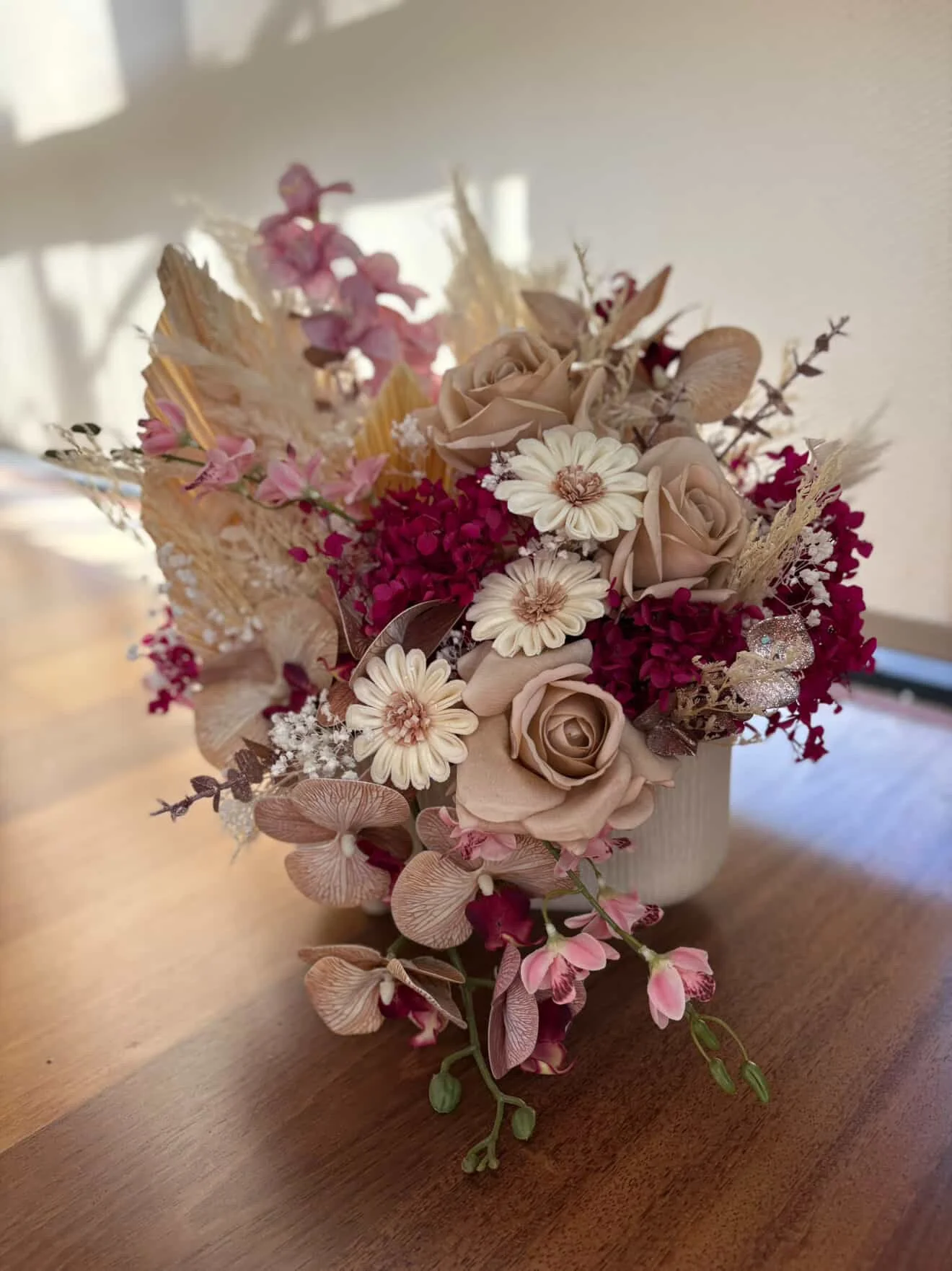 A bouquet of dried flowers with beige, pink, and white blossoms arranged in a white vase on a wooden surface.