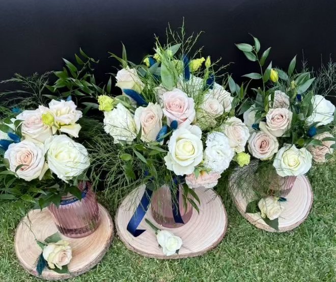 Three floral arrangements in pink glass vases with roses and greenery, placed on wooden slices on grass.