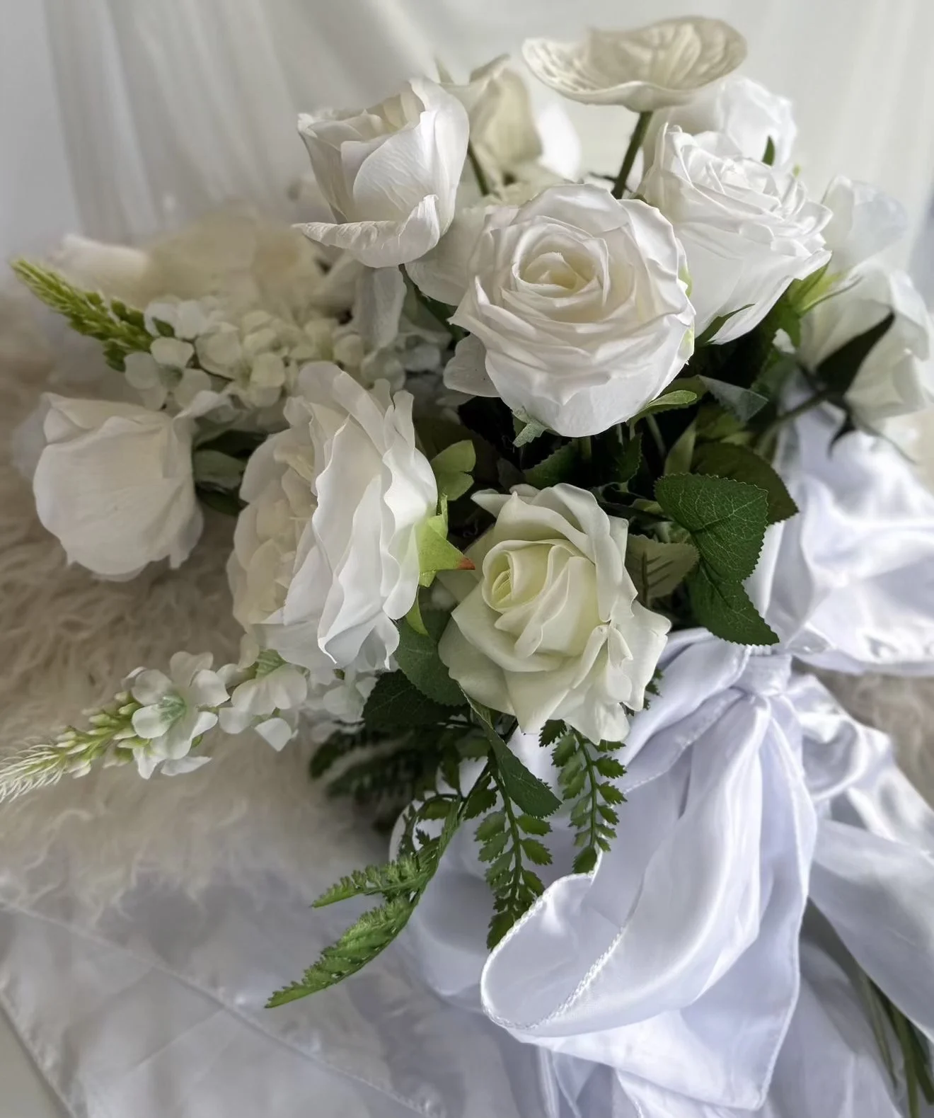 A bouquet of white roses, hydrangeas, and other white flowers with green leaves, wrapped with white ribbon, resting on a soft surface.