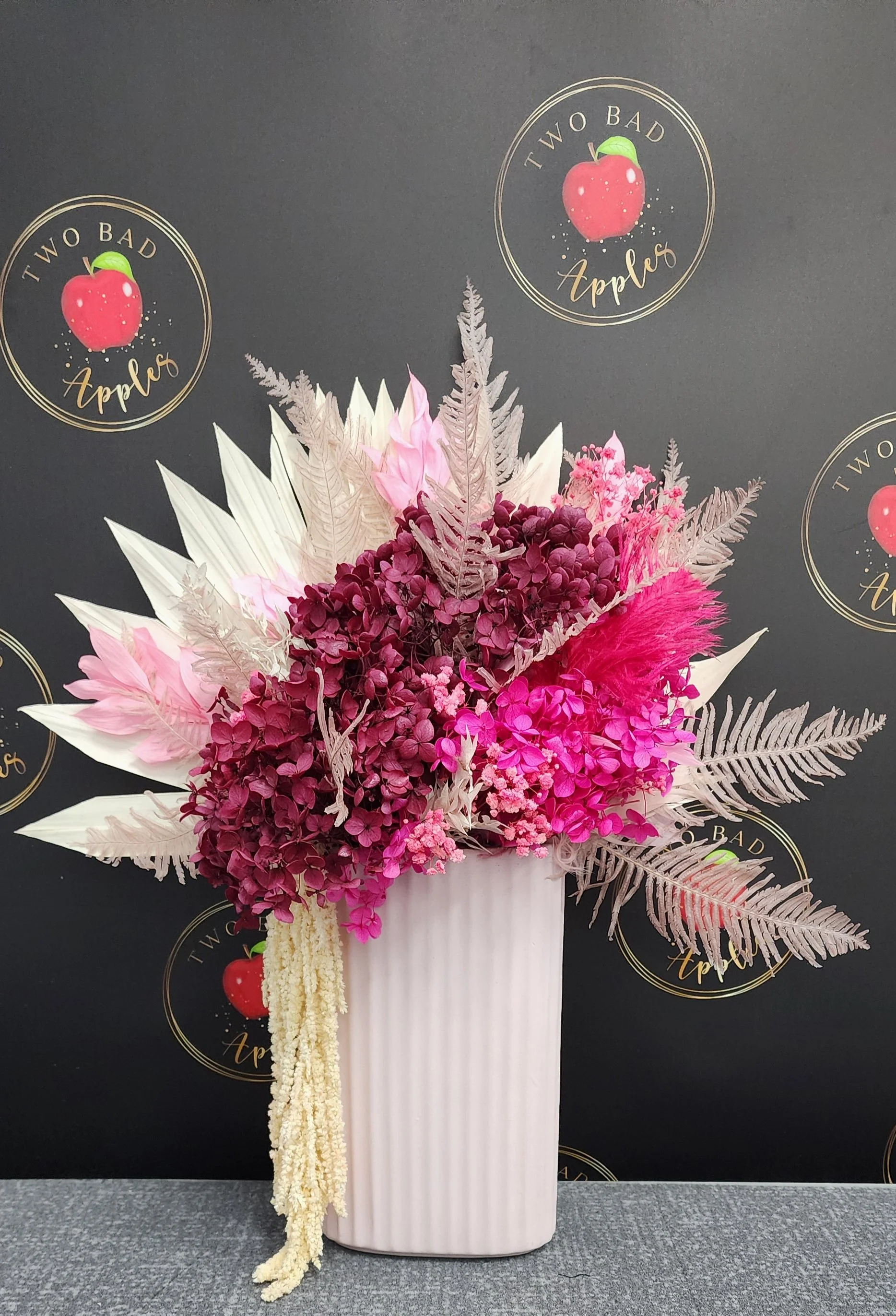 A floral arrangement in a tall, white vase with pink, purple, and white flowers and decorative foliage, set against a black backdrop with gold-colored circular logos that read 'Two Bad Apples' with a red apple illustration.