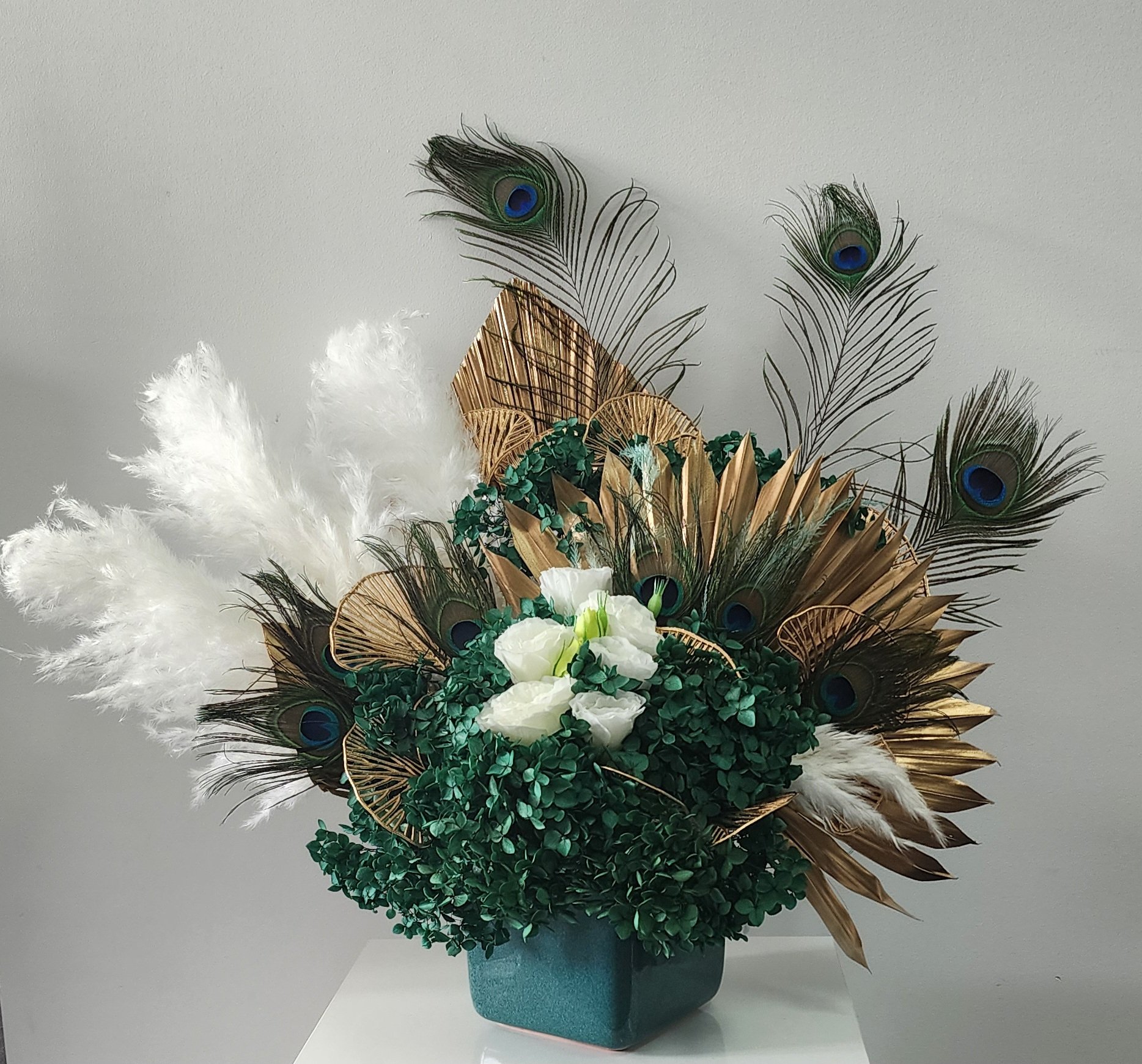 Decorative floral arrangement with white flowers, greenery, peacock feathers, and gold accents in a green vase.