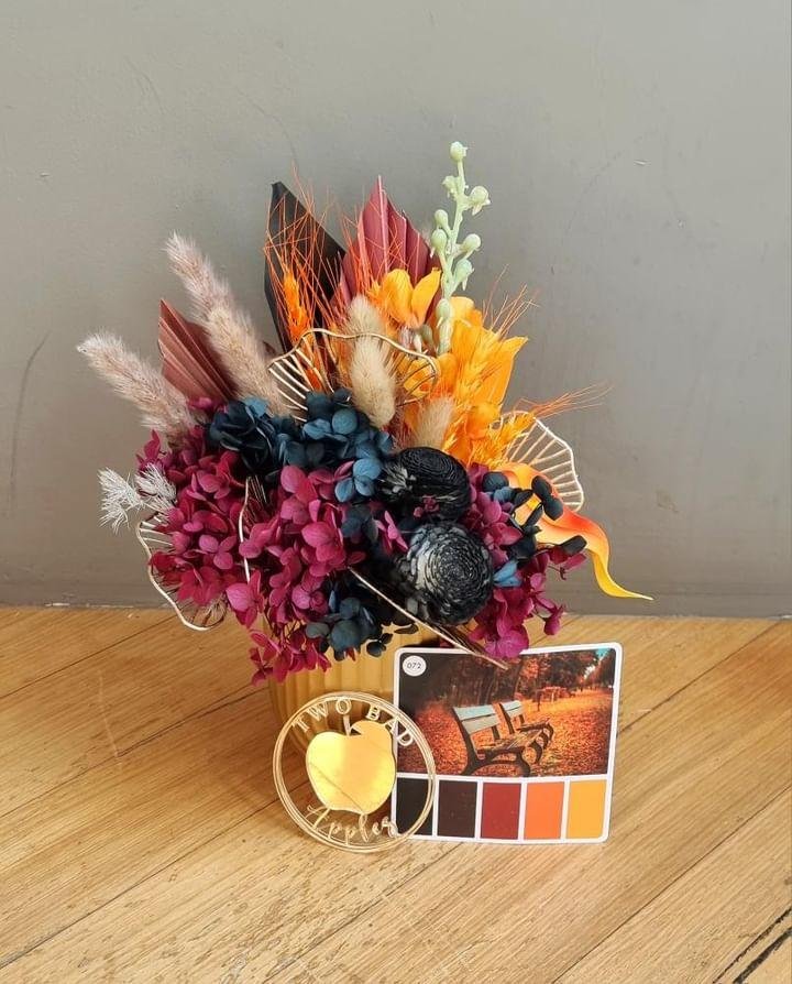 A colorful flower arrangement in a yellow container with pink, orange, yellow, and dark purple flowers, decorative leaves, and dried plants, with a small wood cutout sign reading 'Two Bad' and a color palette card showing shades of orange and red, pl