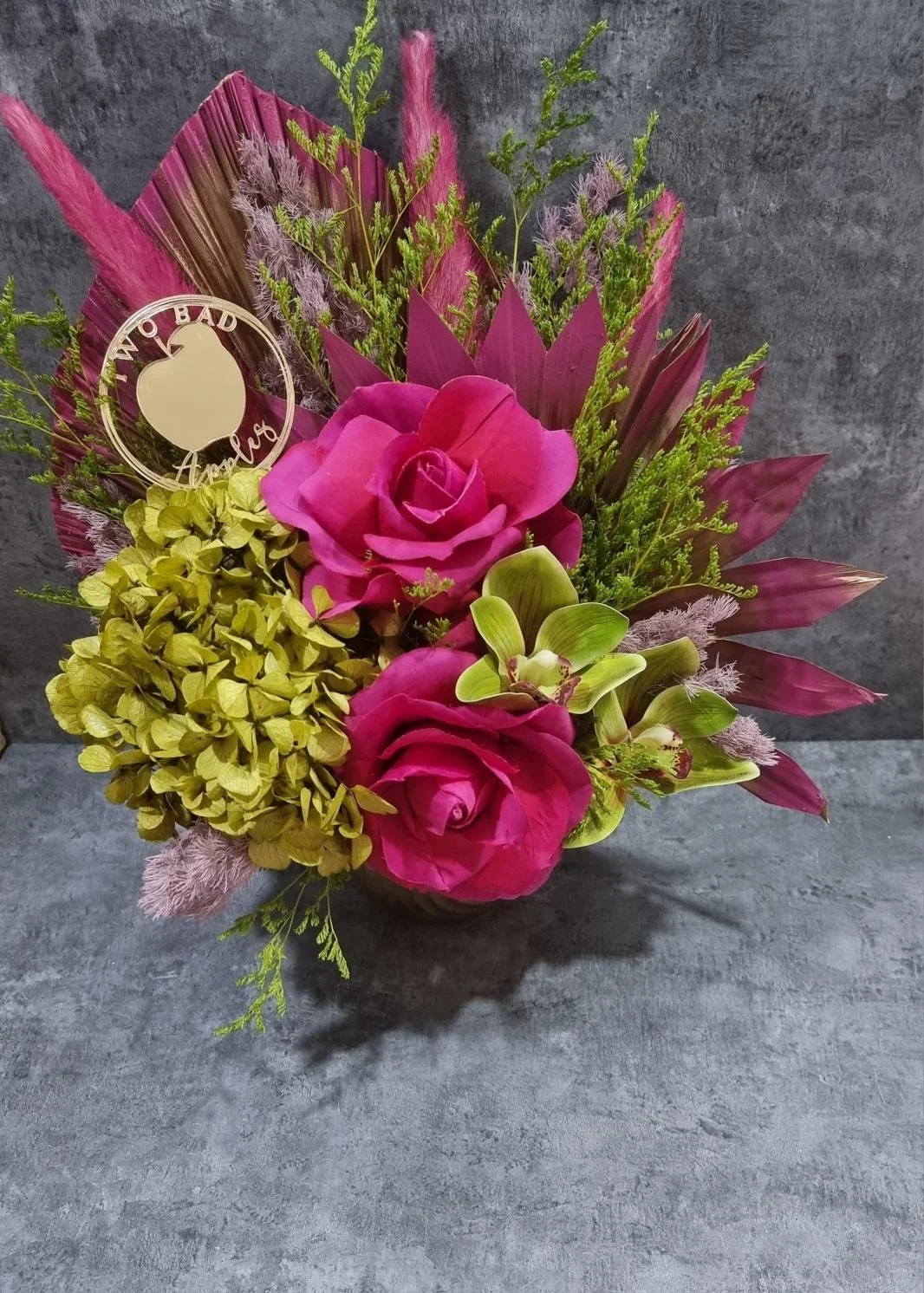 Arrangement of pink roses, green orchids, yellow hydrangeas, and foliage, with a decorative tag reading "I'm not bad, I just have anger" on a gray background.