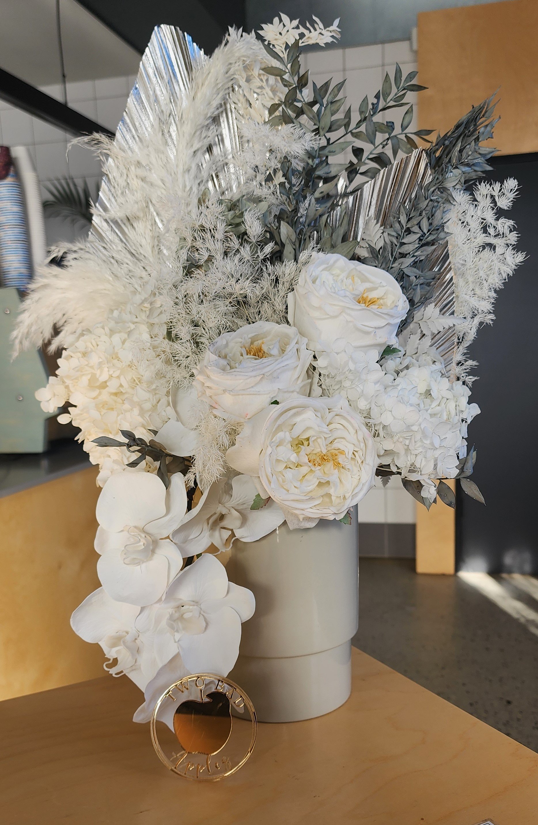 A floral arrangement featuring white roses, hydrangeas, and various white and silver foliage in a white vase, with a gold ring in front of it on a wooden surface.