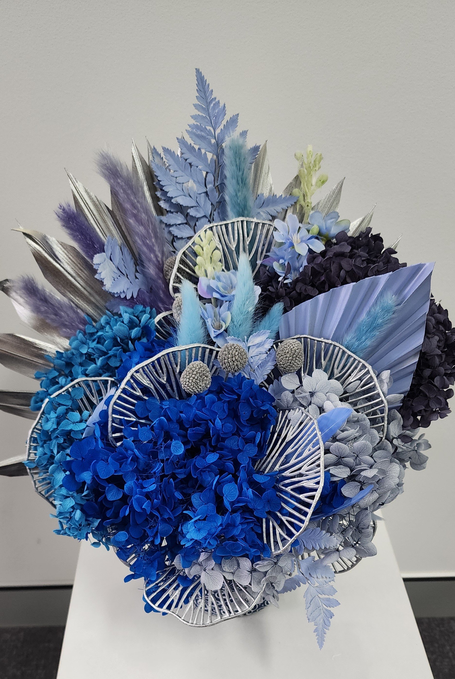 Artificial floral arrangement with blue, purple, silver, and white flowers and foliage in a decorative silver wire basket.