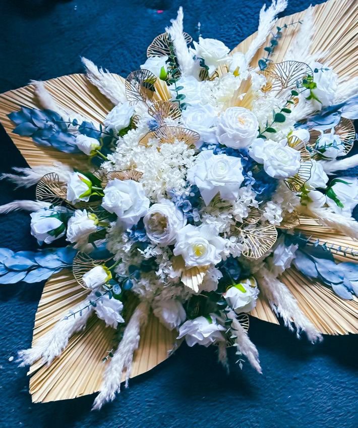 A decorative floral arrangement with white roses, hydrangeas, and other white flowers, accented with gold and blue leaves, set against a dark background.