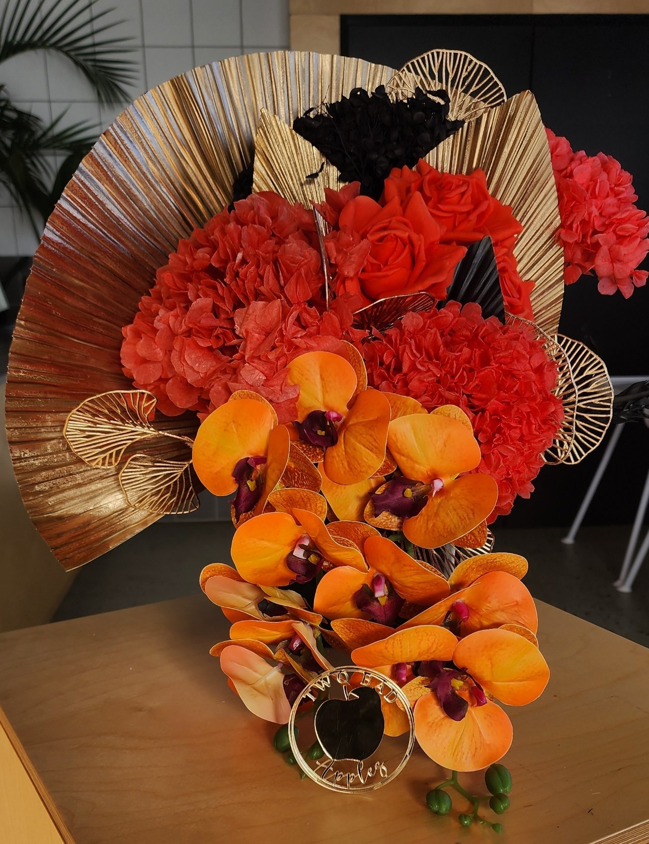 A colorful flower arrangement featuring orange orchids, red roses, pink hydrangeas, gold decorative leaves, and a black circular sign with gold lettering that says 'Thank You'.