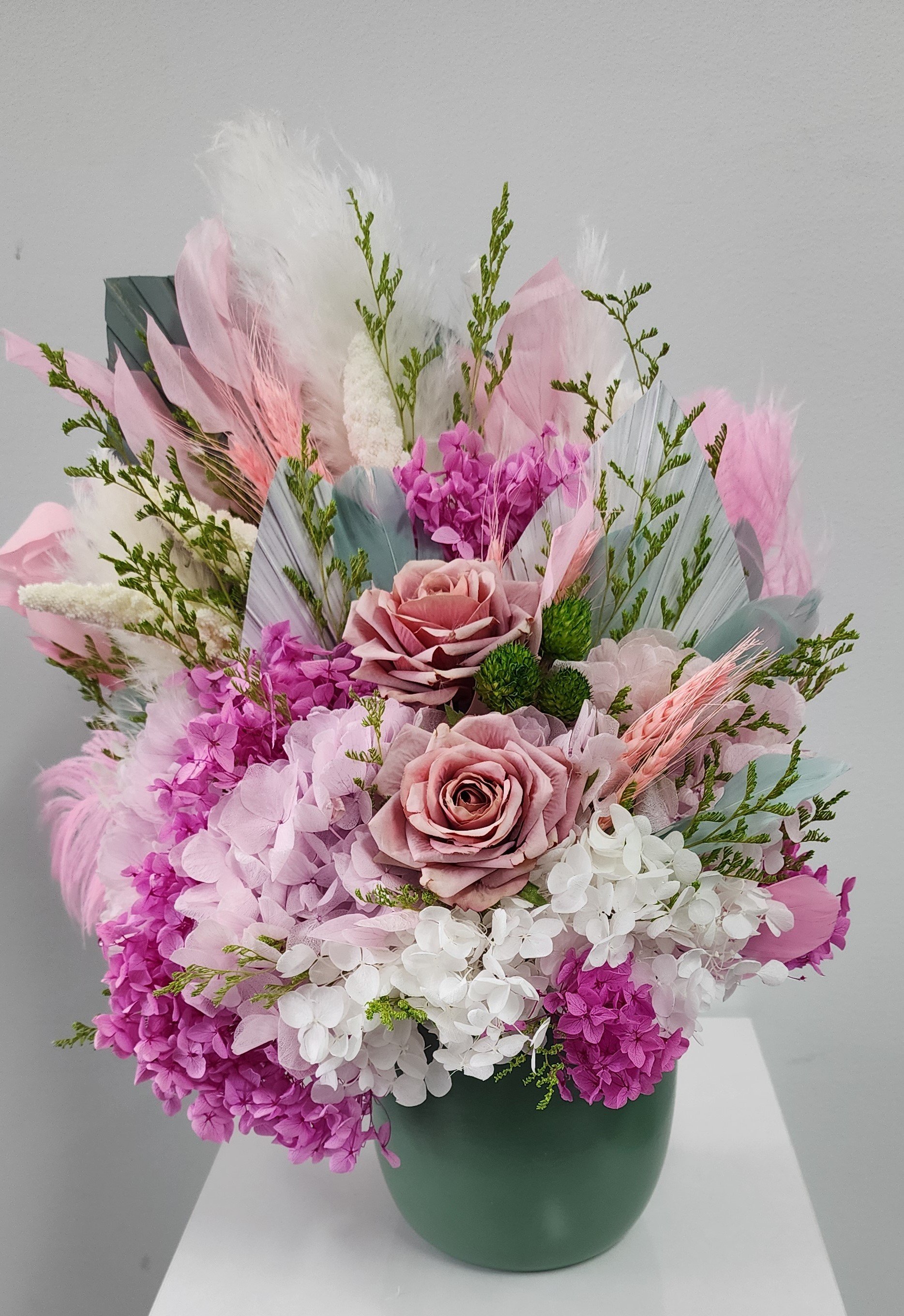 A floral arrangement with pink and white flowers, green leaves, and feathers in a green vase on a white surface.