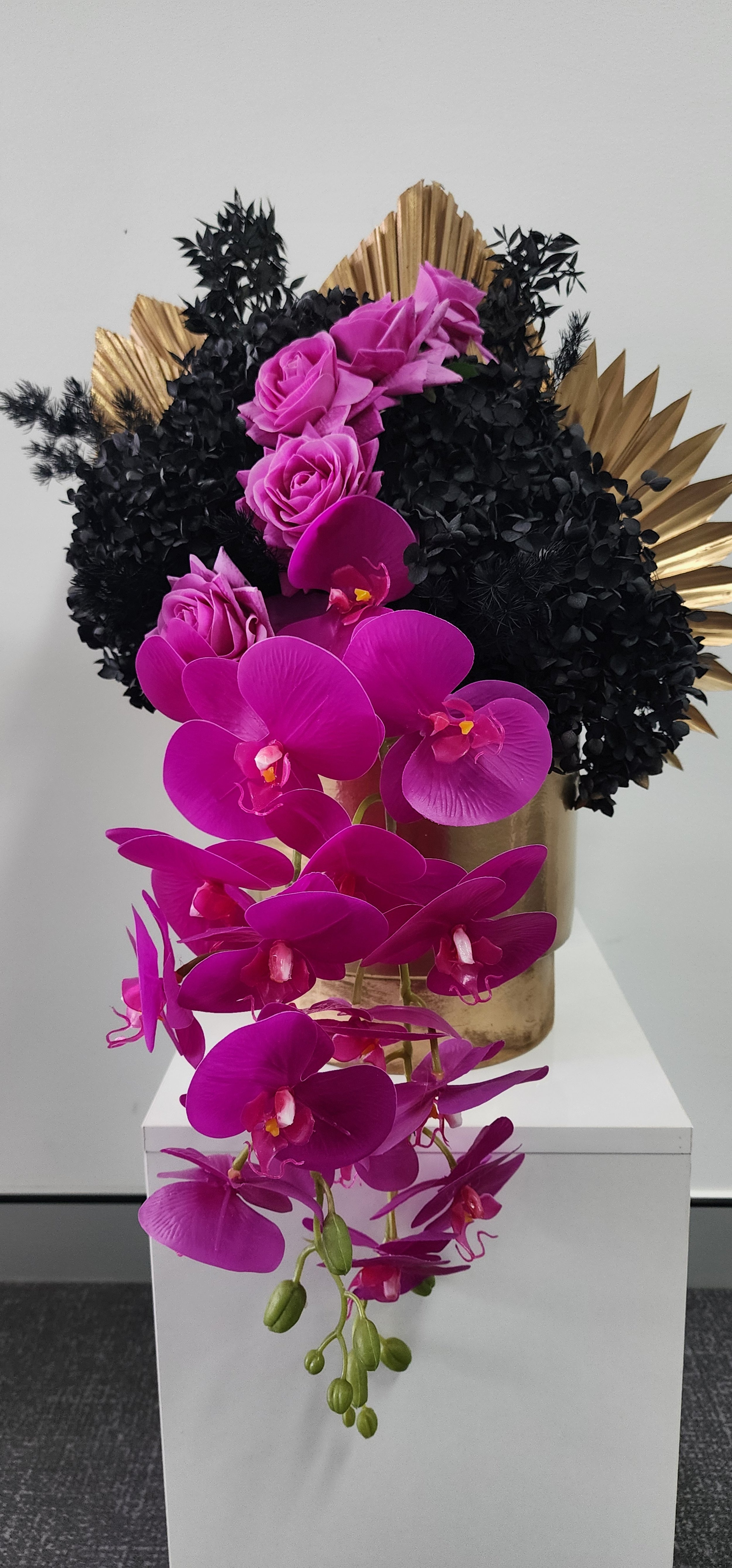 A floral arrangement with pink roses, purple orchids, and black and gold decorative foliage in a gold vase on a white pedestal.
