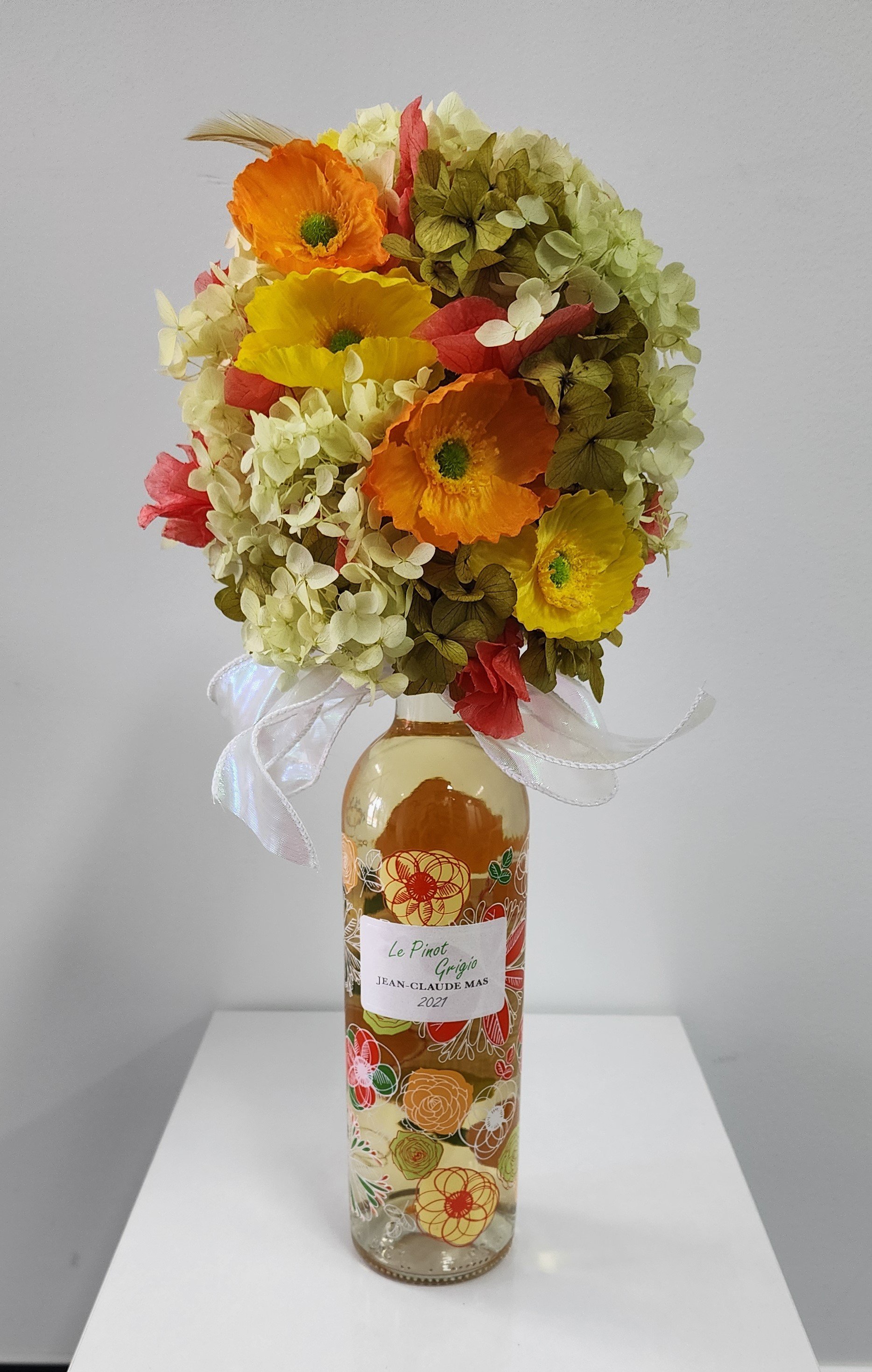 A bouquet of colorful flowers, including yellow, orange, and pink poppies, and cream-colored hydrangeas, in a decorated glass bottle with a ribbon, labeled "Le Pinot Grigio."
