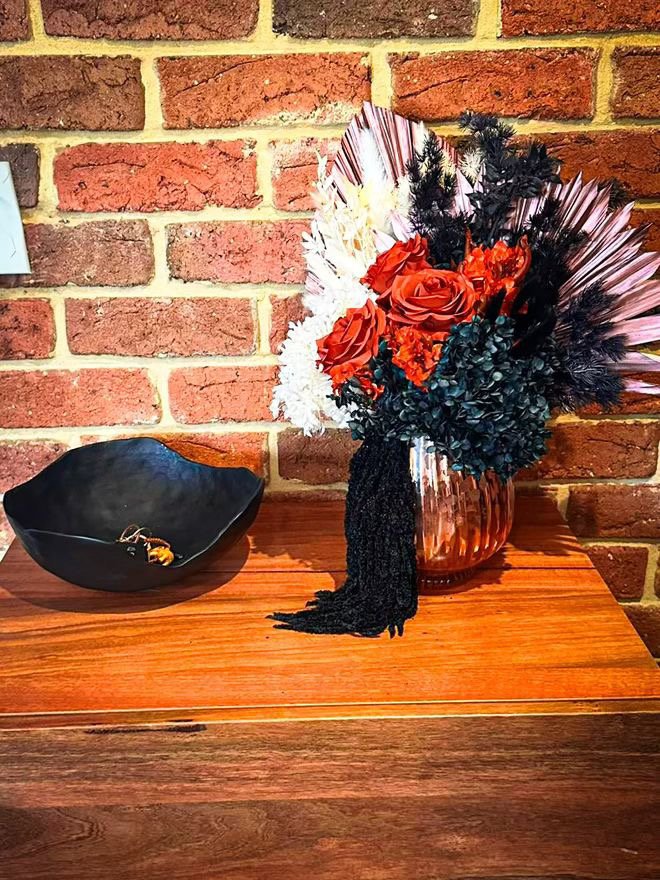 A brick wall background with a wooden surface holding a black bowl containing keys and a decorative flower arrangement in a copper-colored vase with red roses, dark greenery, and dried leaves.