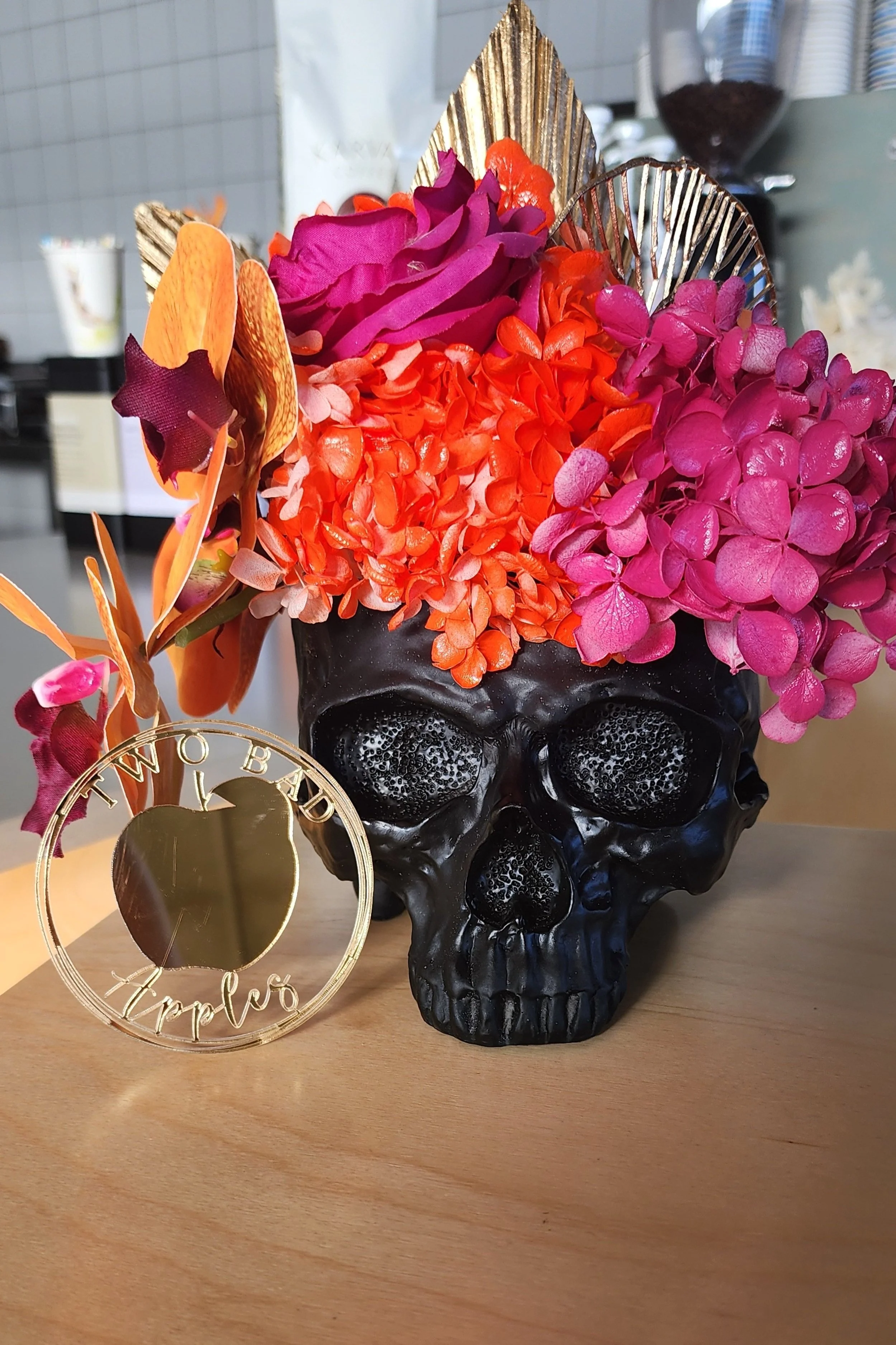 A decorative black skull-shaped vase filled with bright pink, purple, and orange flowers. A gold circular ornament with the word 'Hello' is in the foreground, leaning against the vase. The background includes blurred kitchen items and a coffee grinde