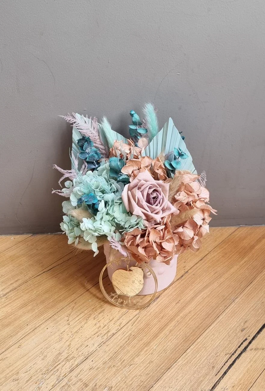 A pastel-colored artificial flower bouquet in a white vase with a gold decorative heart at the front, placed on a wooden surface against a gray wall.