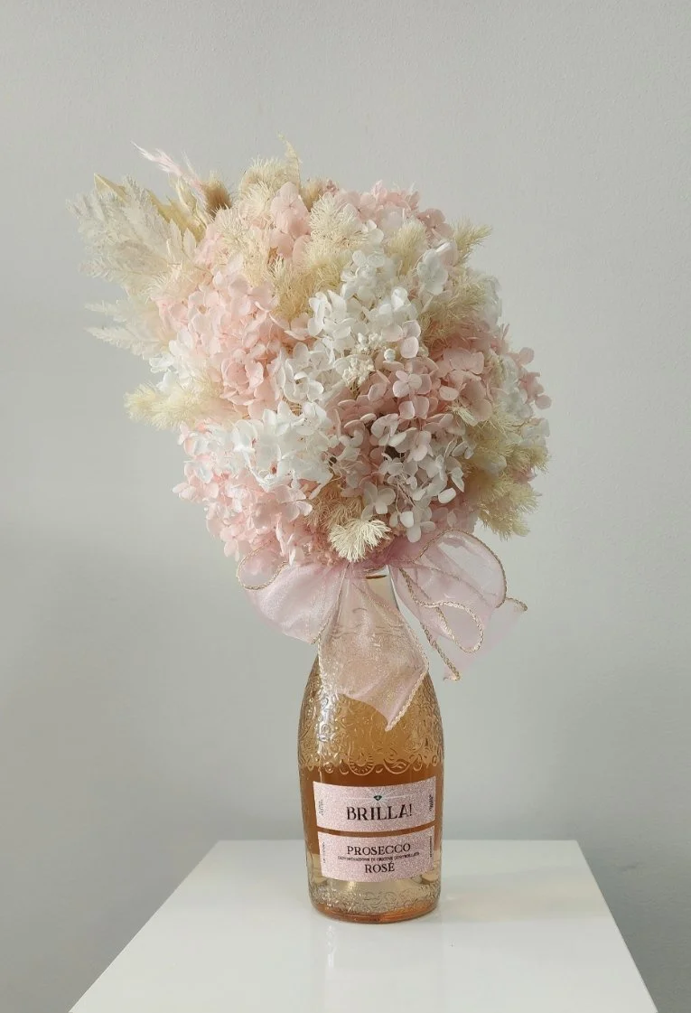 A bouquet of pink and white hydrangeas and dried flowers in a bottle-shaped vase with a pink ribbon, placed on a white surface.