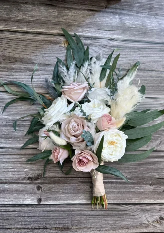 A bouquet of flowers with pink and white roses, green leaves, and dried grasses on a wooden surface.