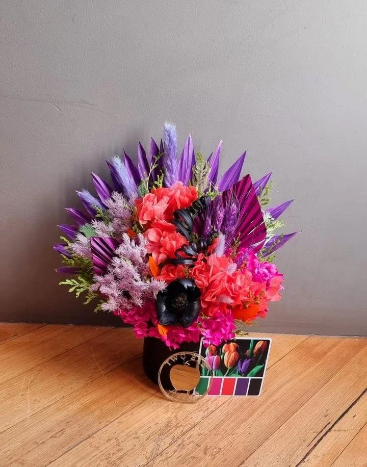 Colorful floral arrangement in a black vase with various purple, pink, red, and black flowers, positioned on a wooden surface against a gray wall.