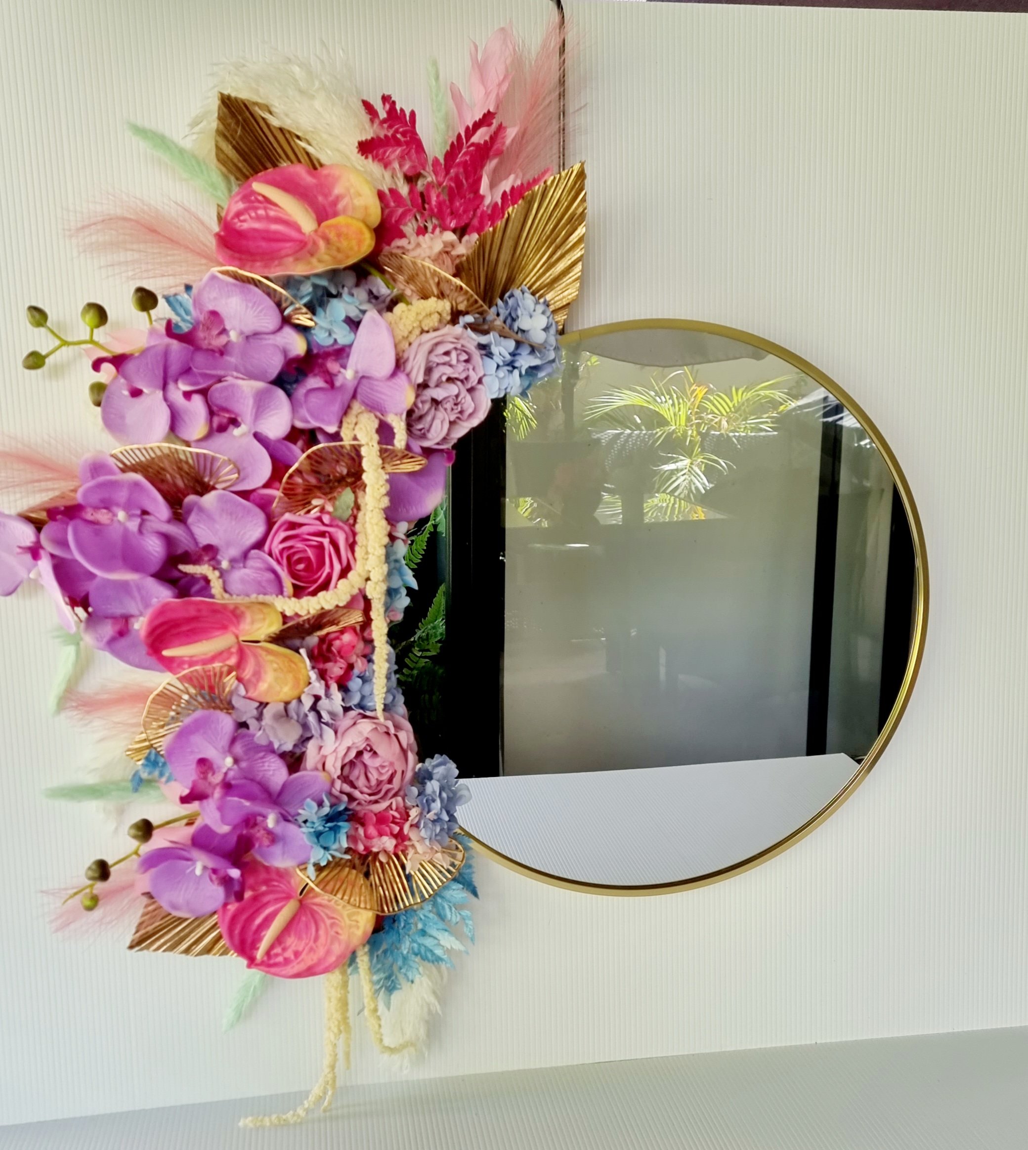 A colorful floral arrangement with pink, purple, and blue flowers, gold decorative elements, and various foliage, displayed on a wall with a reflection of plants in the mirror nearby.