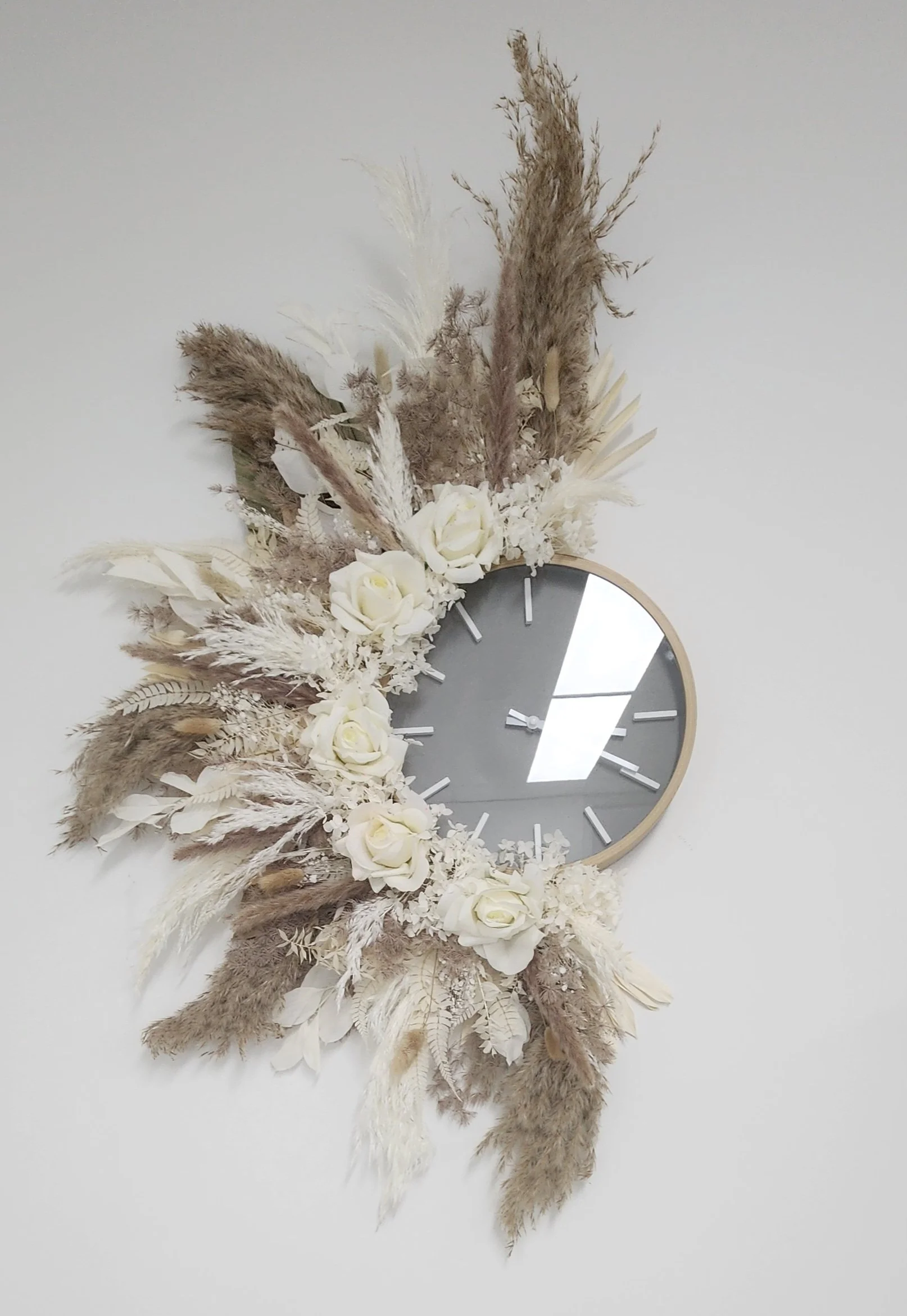 A round wall clock surrounded by a floral arrangement of white roses, dried pampas grass, and other dried flowers on a white wall.