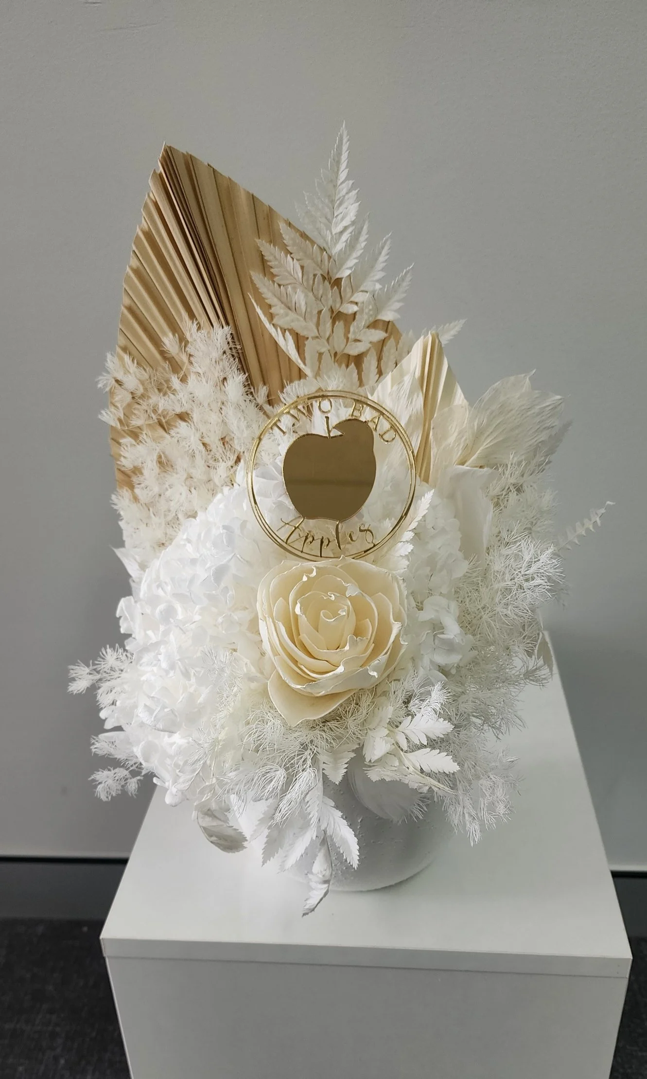 A white floral arrangement in a white vase featuring white roses, dried palm leaves, wheat, and various white foliage, with a round gold sign that reads 'The Best Wishes' attached to the arrangement.
