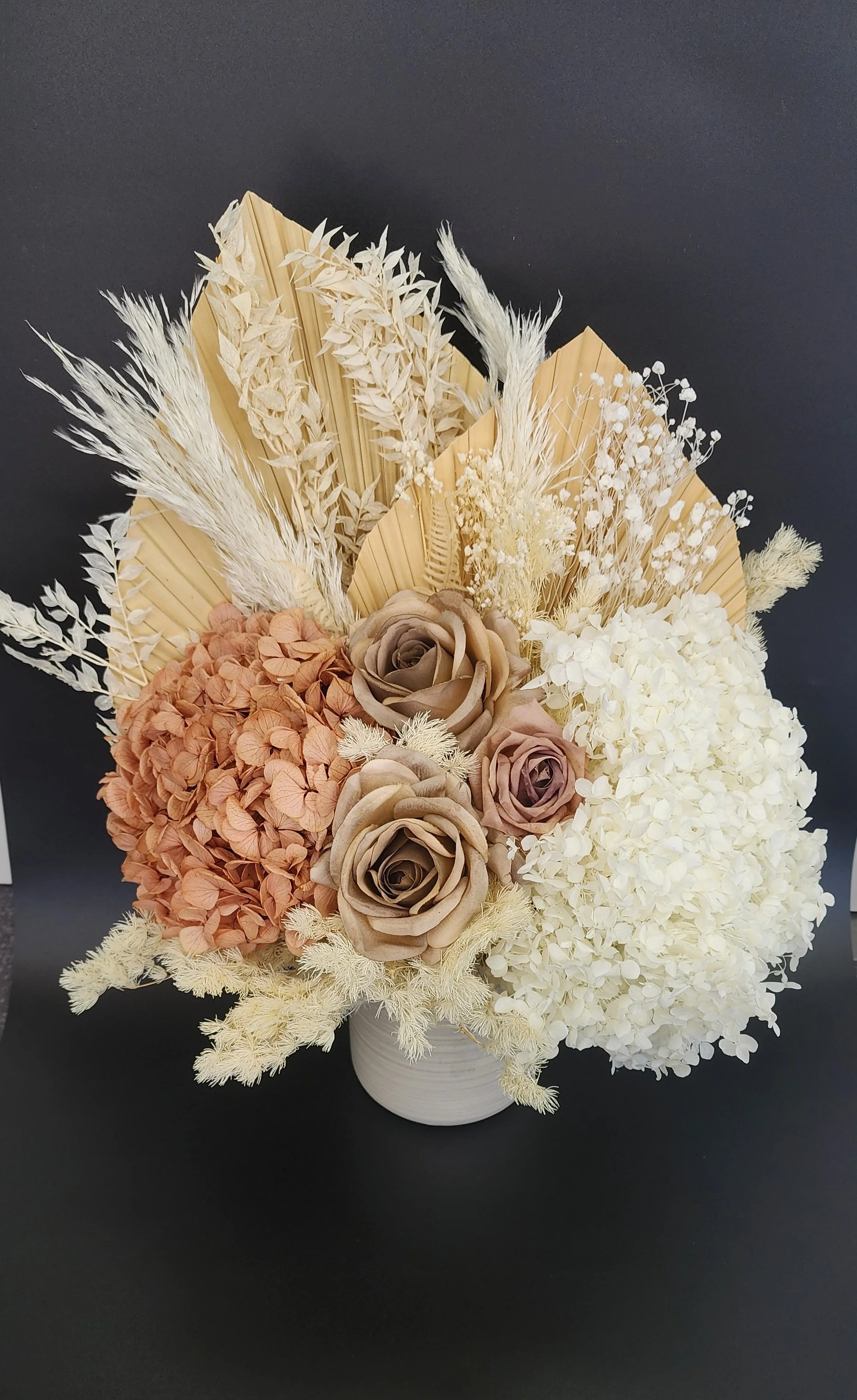Arrangement of dried flowers in neutral tones with cream, beige, and light brown colors, featuring roses, hydrangeas, pampas grass, and palm leaves in a white vase against a black background.
