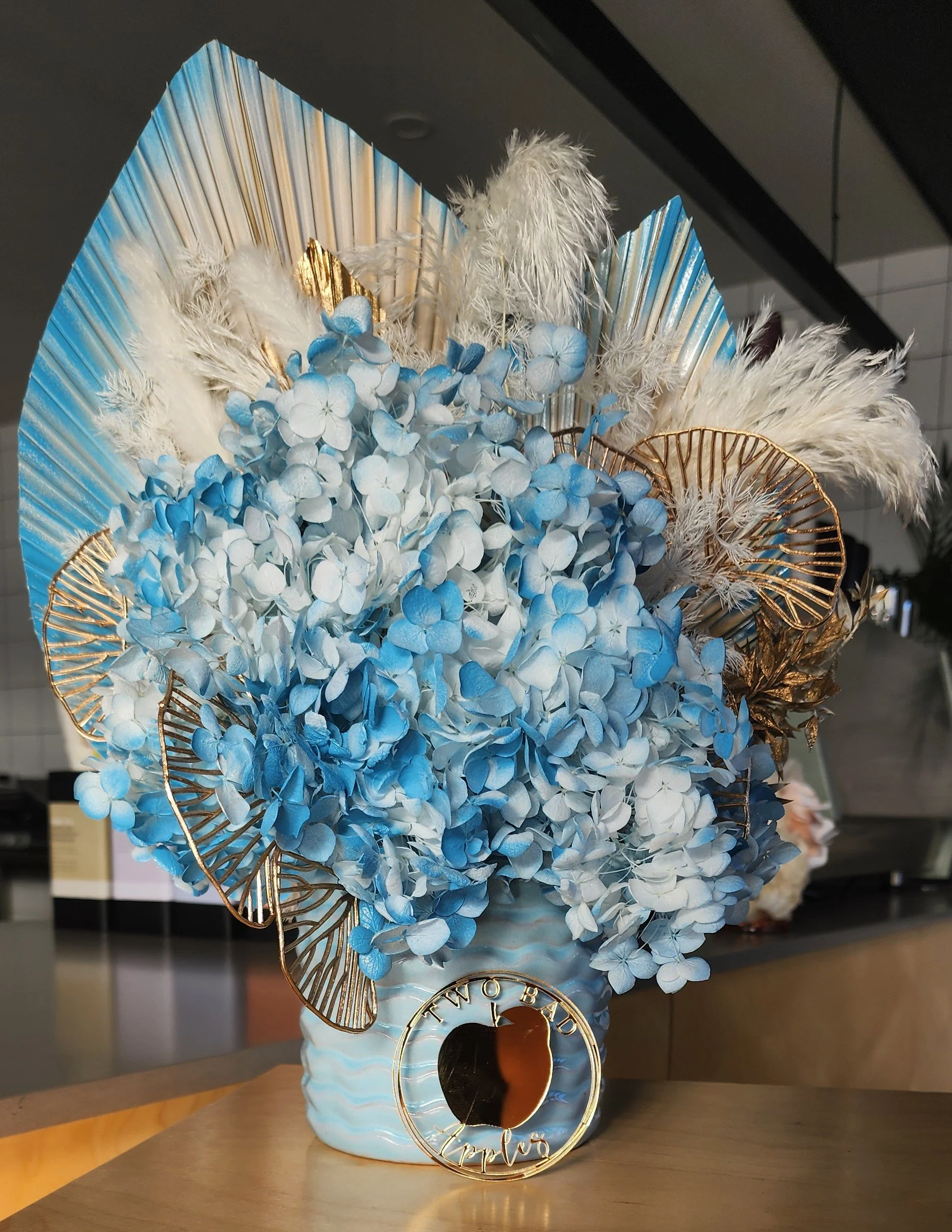 Decorative arrangement with blue and white hydrangeas, white pampas grass, gold accents, and a ceramic vase, with a circular gold sign reading 'Love' in cursive.