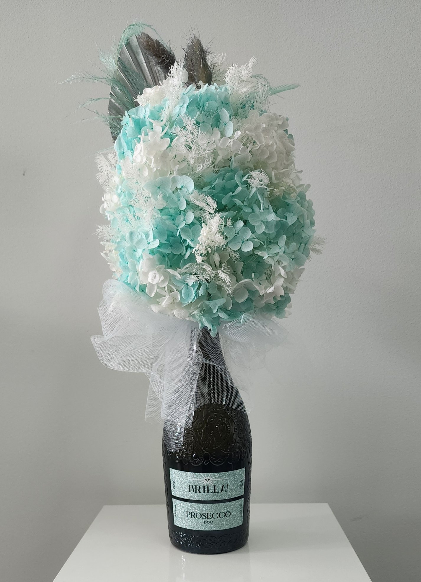 A bouquet made of white and light blue hydrangeas with feathers, arranged in a dark wine bottle labeled 'Brilla! Prosecco DOC', wrapped with white tulle, set on a white surface against a plain light gray wall.