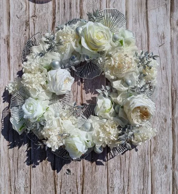 White floral wreath with roses, hydrangeas, and decorative silver leaves, placed on a wooden surface.