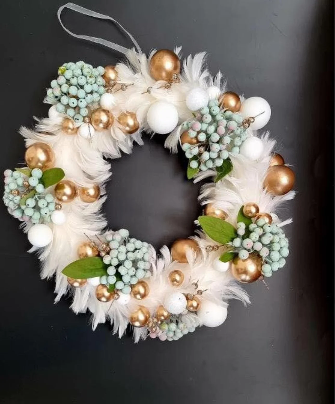 Festive wreath made of white feathers, green berries, white and gold ornaments, and green leaves on a black background.