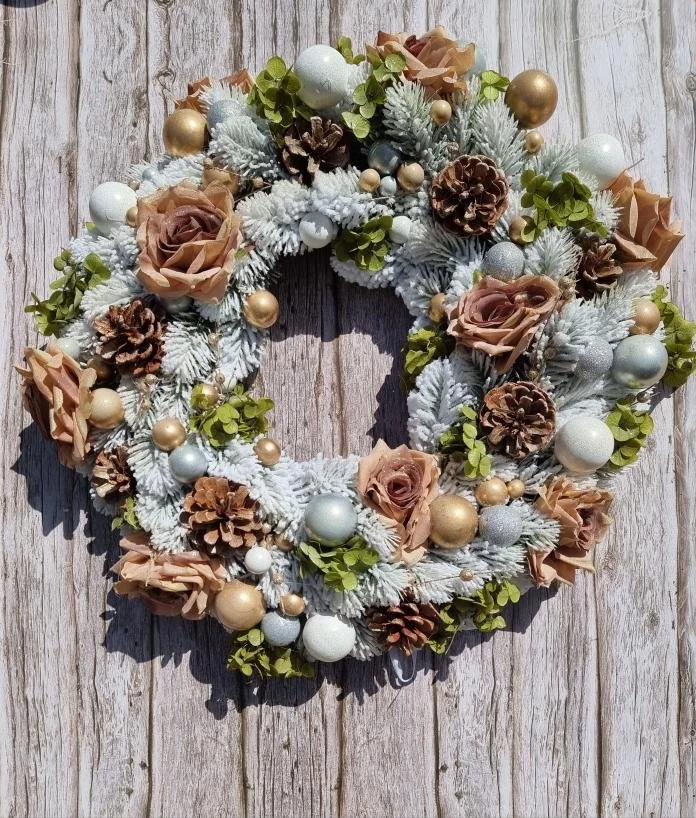 Decorative Christmas wreath with pinecones, gold and silver ornaments, beige roses, green foliage, and white frosted branches, hanging on a wooden surface.