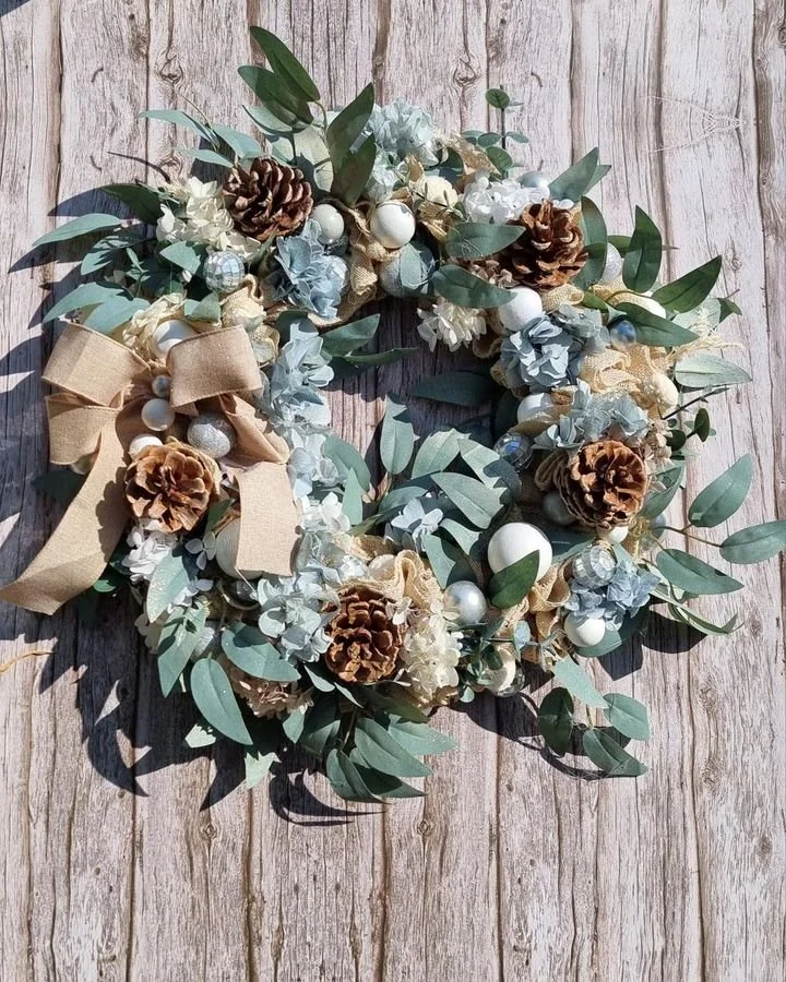 A decorative wreath adorned with pinecones, white and silver ornaments, cream-colored ribbons, and green foliage, placed on a weathered wooden surface.