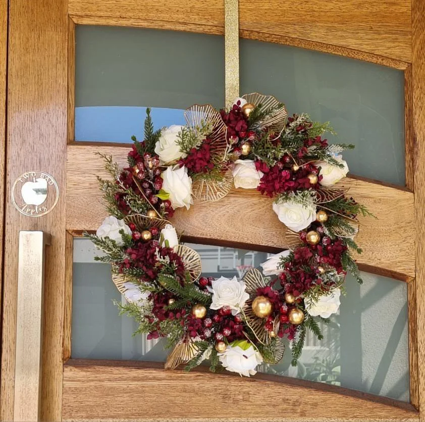 Christmas wreath with white roses, red berries, gold ornaments, and greenery hanging on a wooden door.